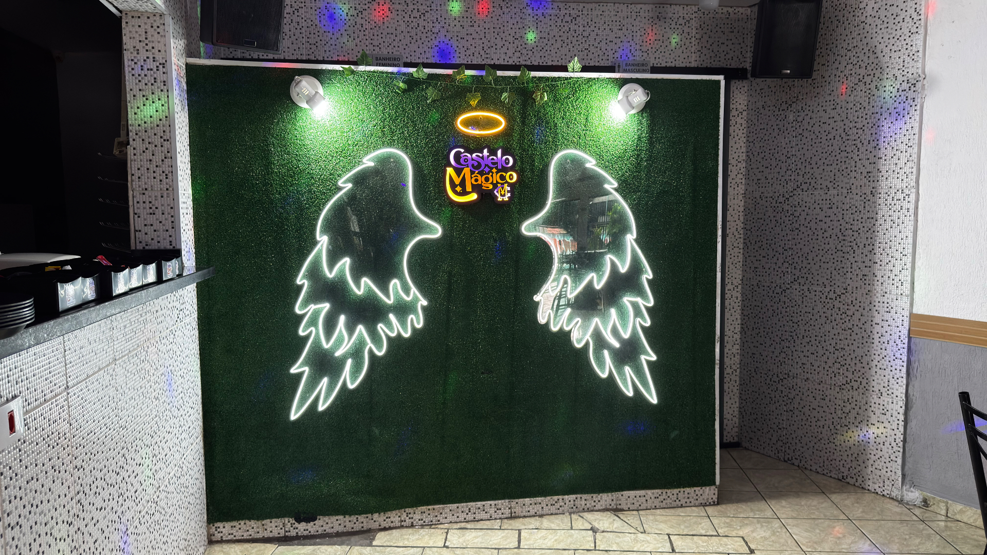Espaço Asas Mágicas do Castelo Mágico Buffet Infantil, com parede verde e letreiros de asas em neon ao redor do logo do buffet